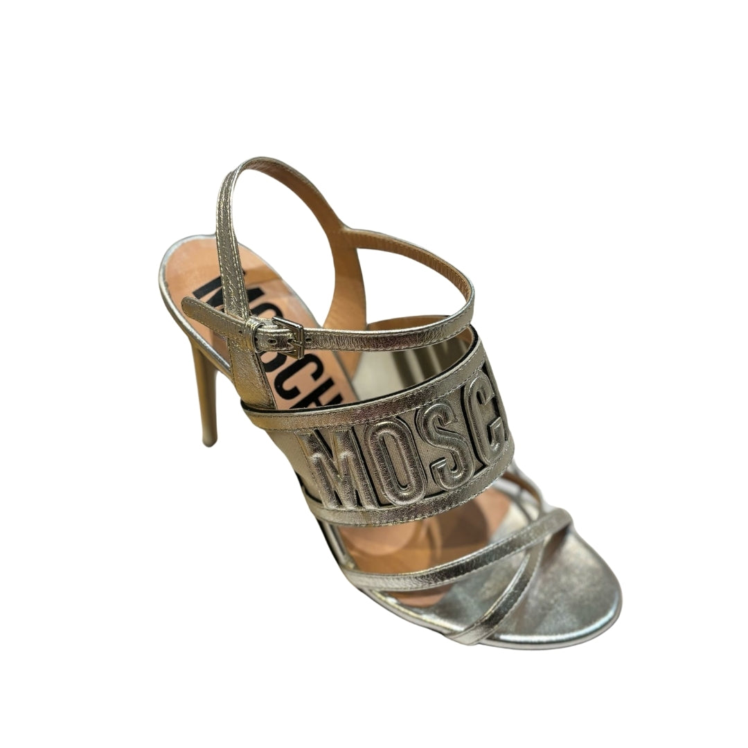 Moschino silver metallic heeled sandals featuring an open toe, stiletto heel, ankle strap with buckle, and a wide front strap with raised Moschino logo lettering, available at Lestyle Boutique 136 Little Collins St.