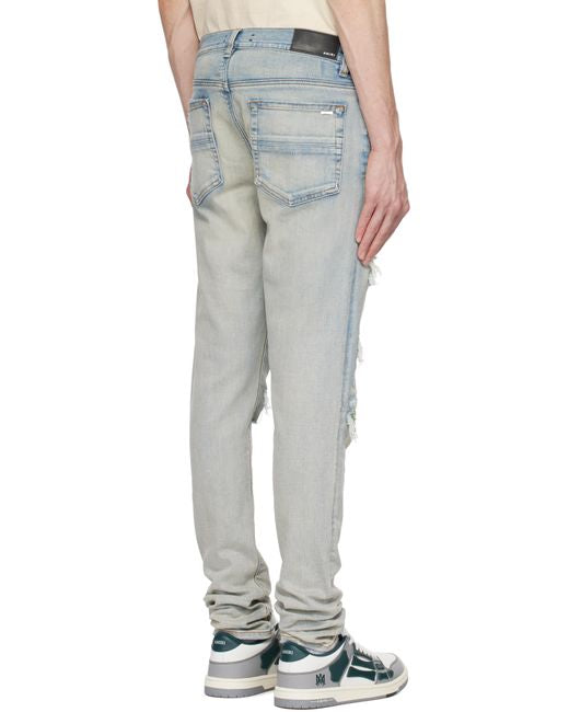 A pair of antiqued indigo stretch skinny jeans by AMIRI, available at Lestyle Boutique. These jeans feature a stonewashed and distressed finish with repaired rips, belt loops, and a contrasting logo label for a contemporary look.