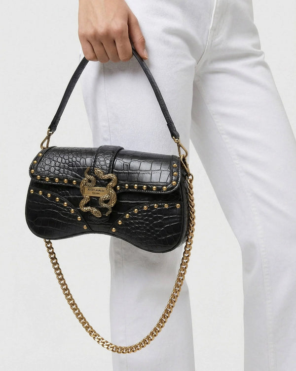 Black handbag with gold chain and studded details held by a person wearing white pants.