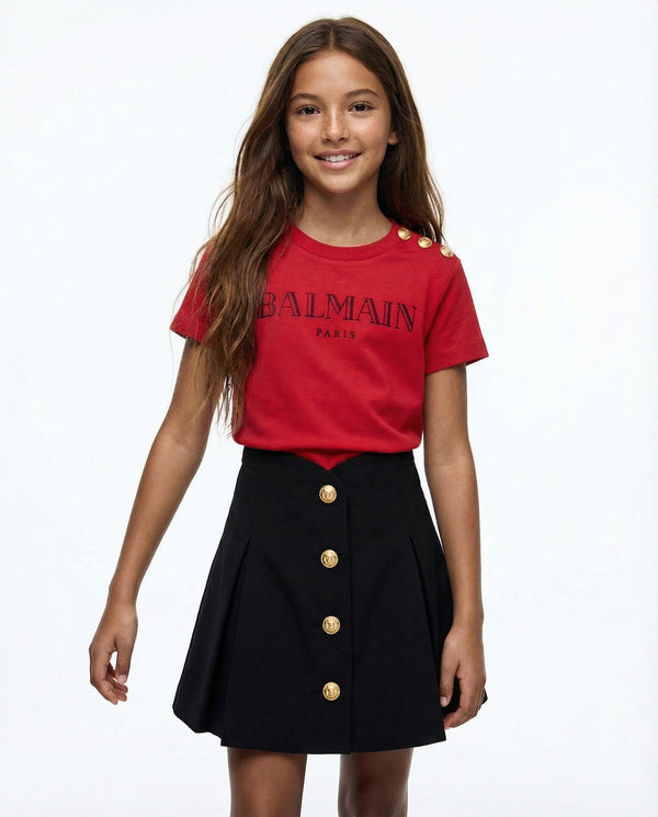 Young girl wearing a red Balmain t-shirt and black skirt with gold buttons on a white background