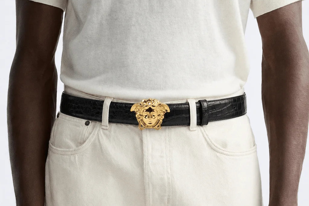 Person wearing a black versace belt with a gold buckle on white pants against a light background