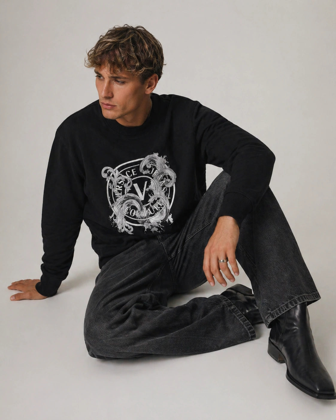 Person wearing a black Versace Jeans Couture sweatshirt with a logo, sitting on the floor against a plain background